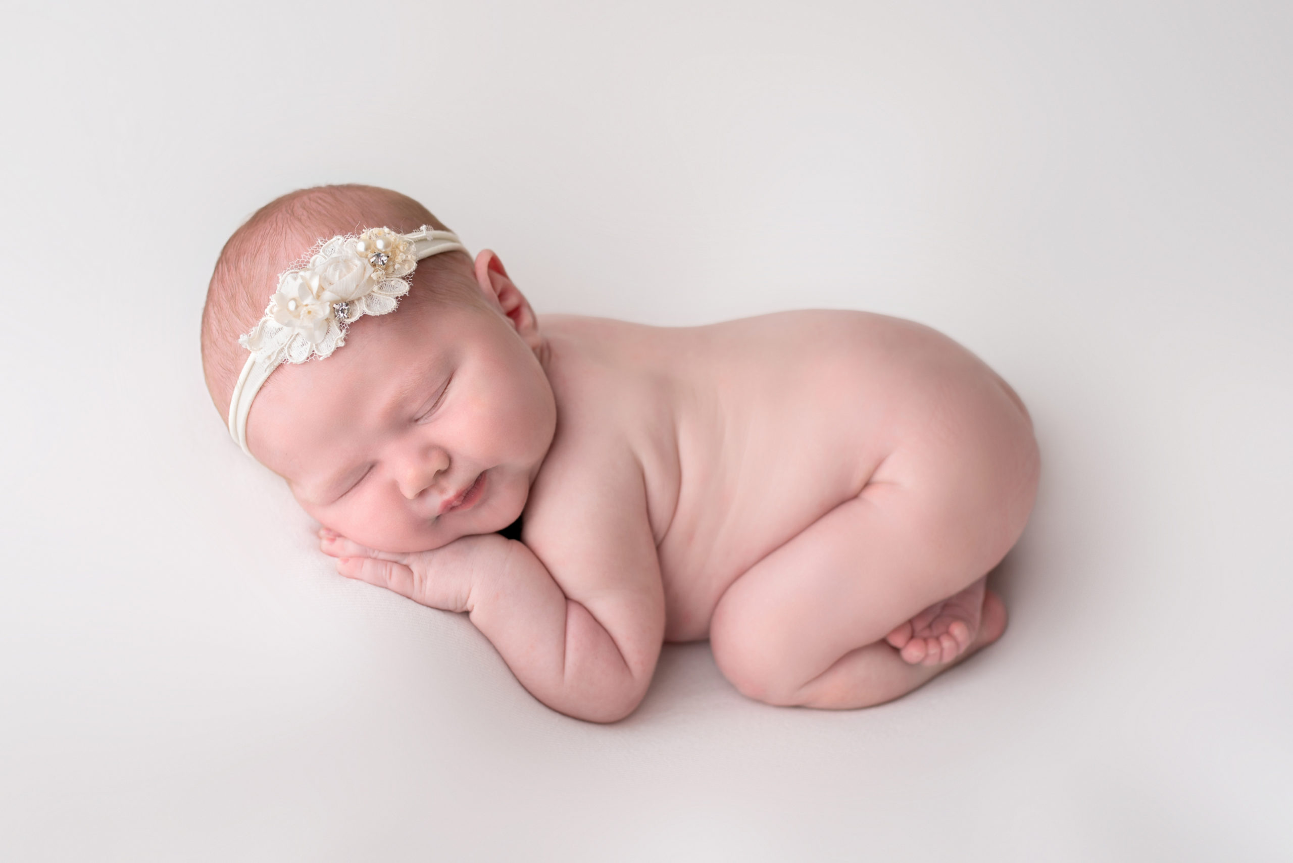 newborn baby girl with a white flower headband laying on a white backdrop Houston Baby Stores