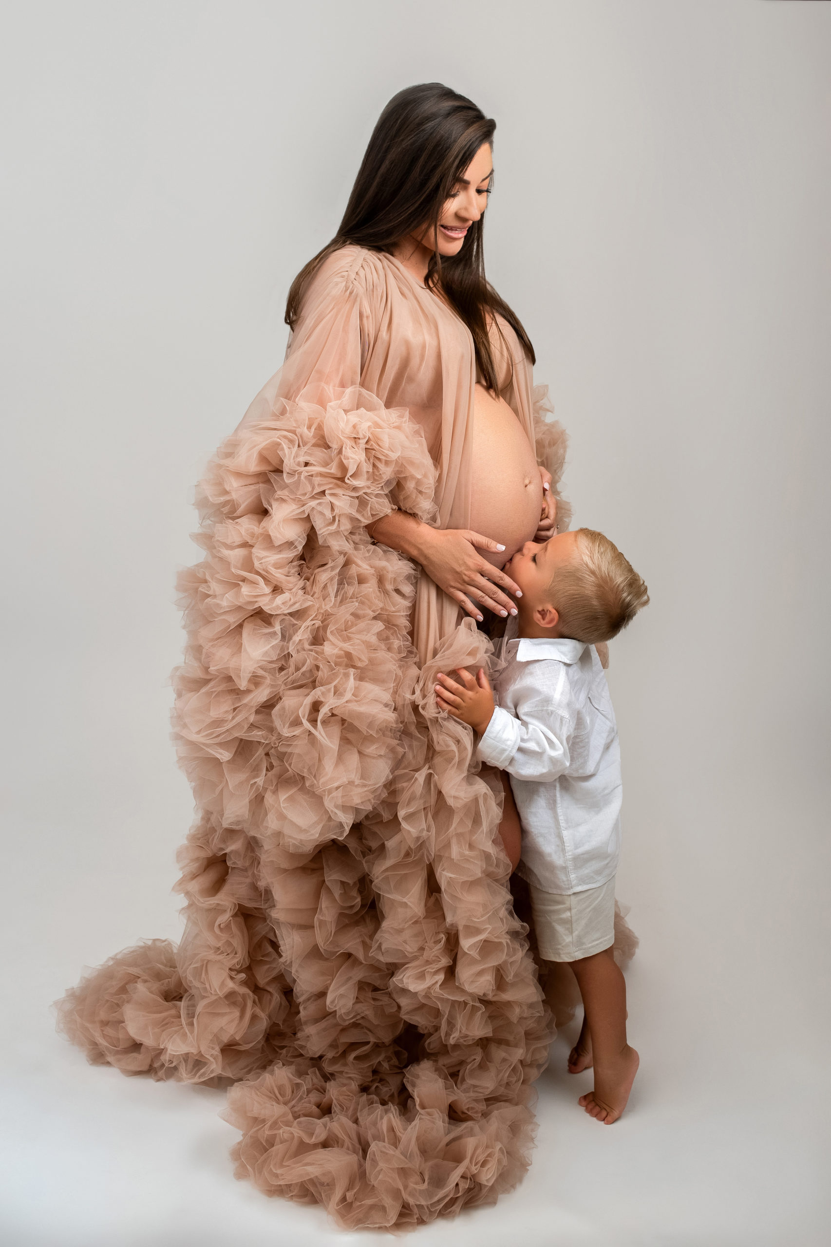 mom to be in neutral tulle gown with son kissing her bump Houston Nanny Agency