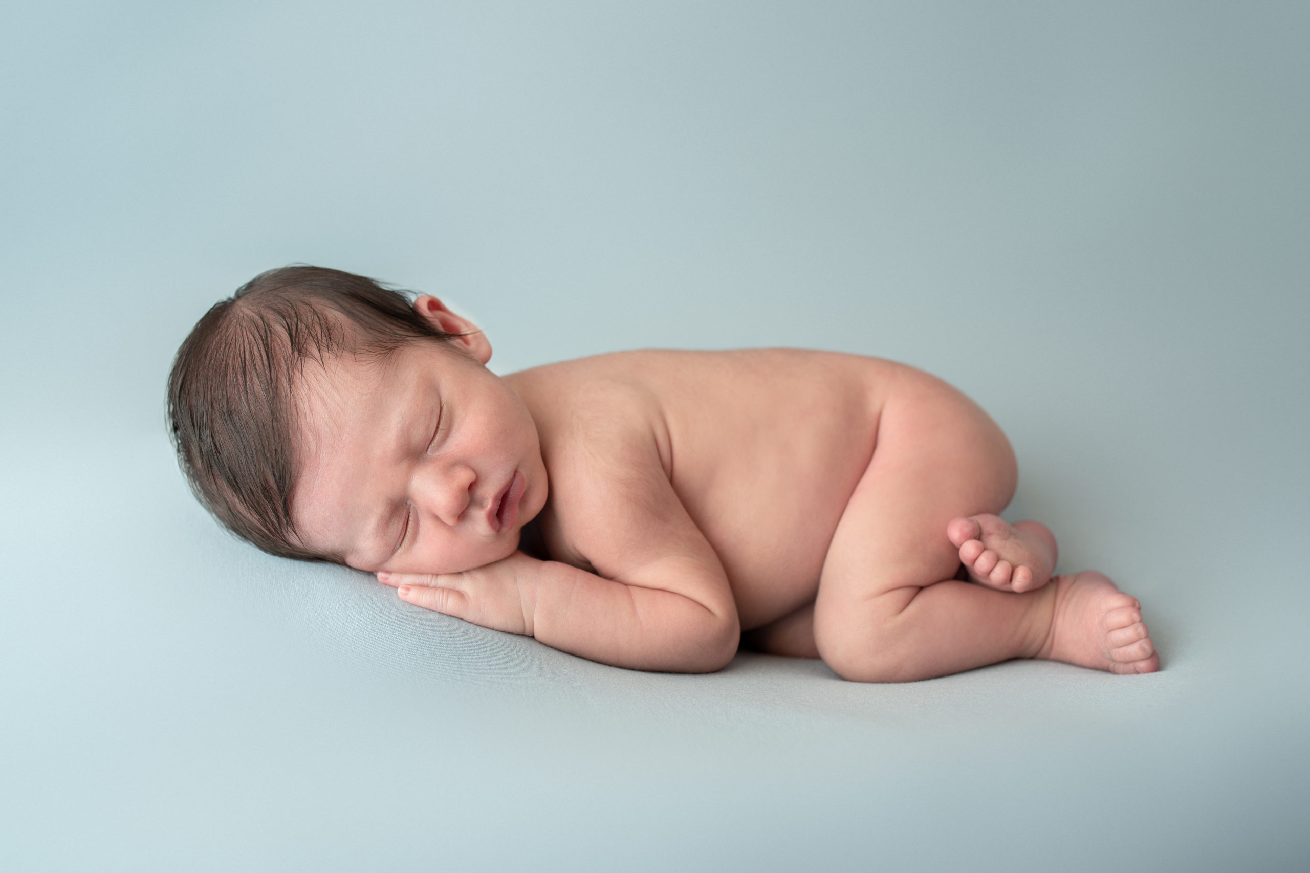 newborn baby without clothes sleeping on it's hands on a blue backdrop