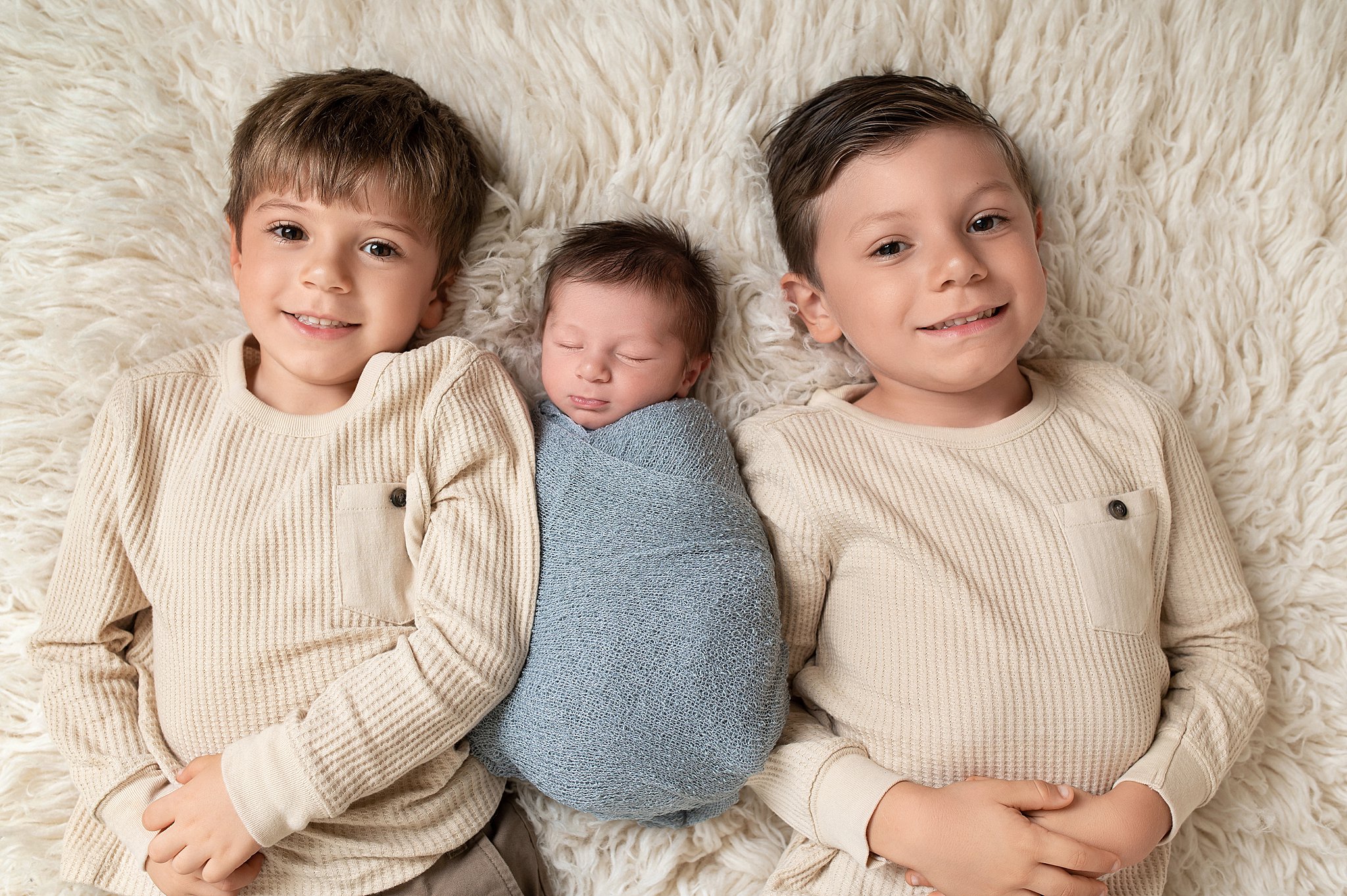 Two brothers wearing matching tan shirts lay on a furry blanket with sleeping newborn sibling Janie and Jack Houston