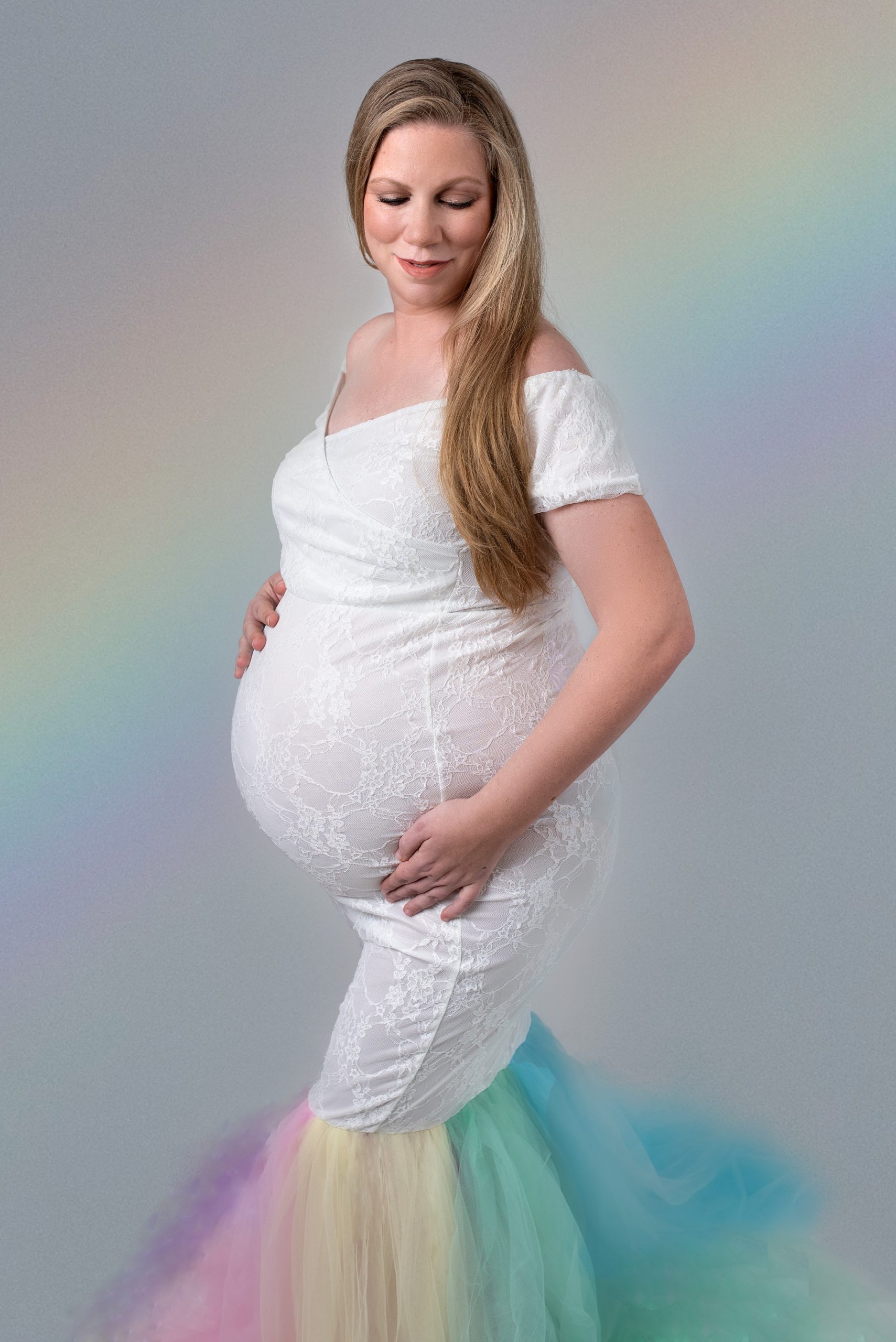 Woman in a rainbow maternity gown stands holding her bump in a studio